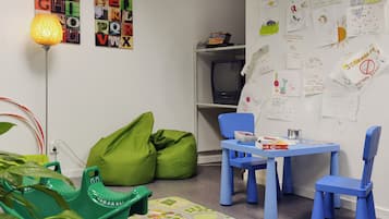 Un cuarto de juegos infantil con una mesa azul, pufs verdes, carteles del alfabeto y un televisor sobre una estantería.
