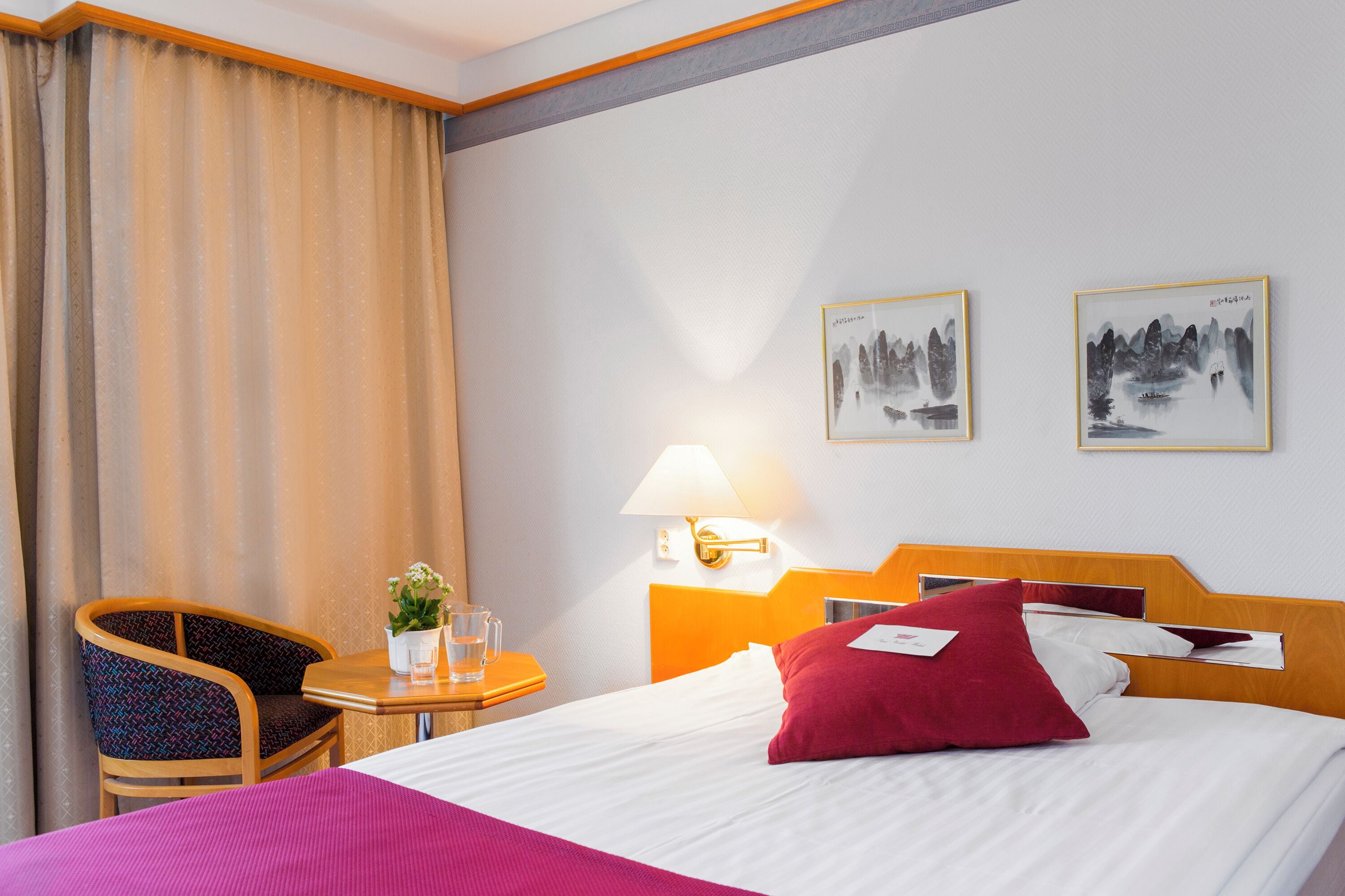 A hotel room with a bed, a chair, a small table with a plant and a glass, and two wall-mounted paintings.