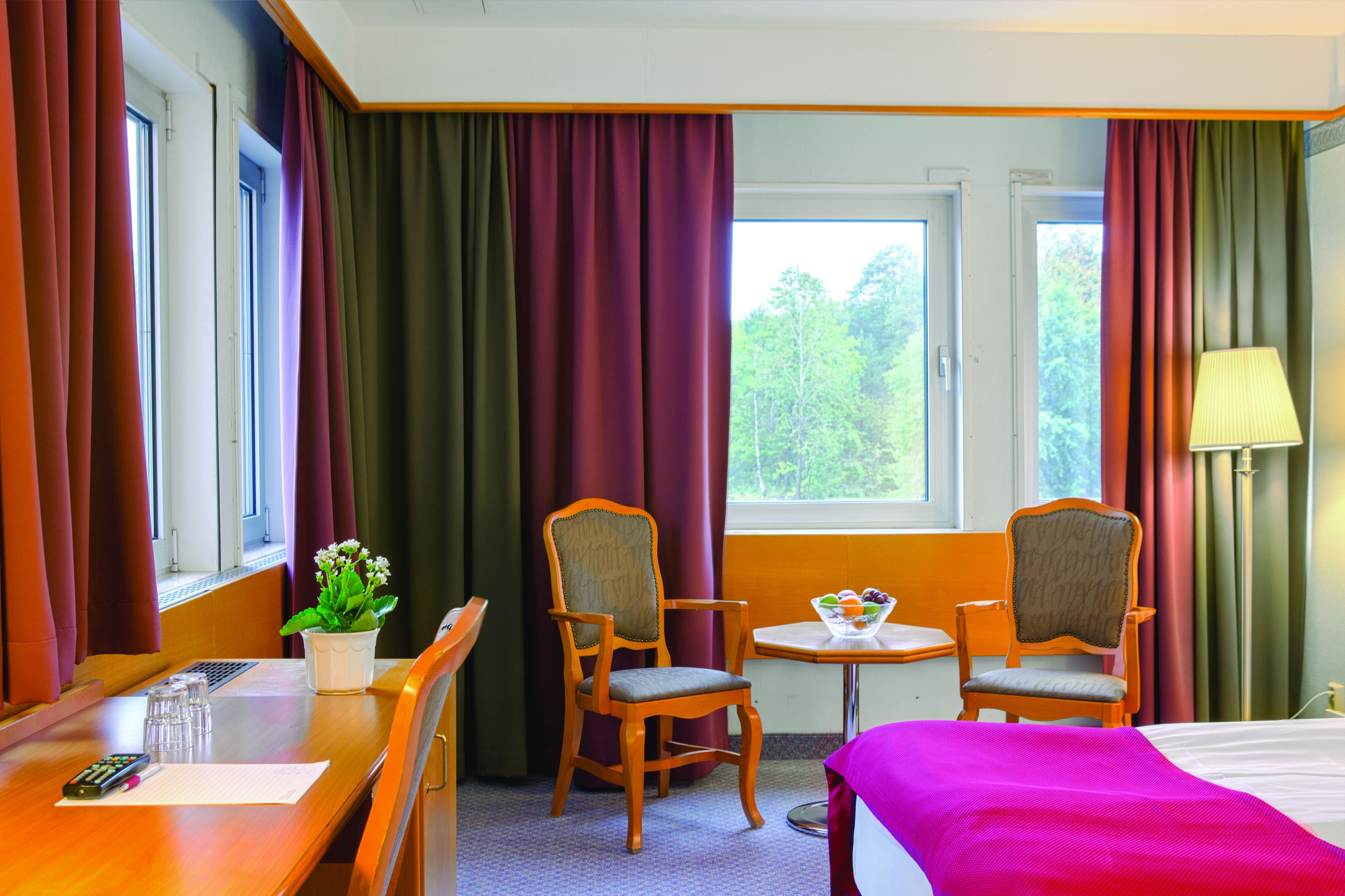 A hotel room with two wooden chairs, a small table with a bowl of fruit, a bed with a pink blanket, and a window with curtains.