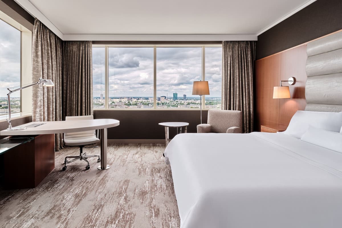 Ein Hotelzimmer mit einem großen Bett, einem Schreibtisch, einem Sessel und Blick auf die Stadt durch große Fenster.