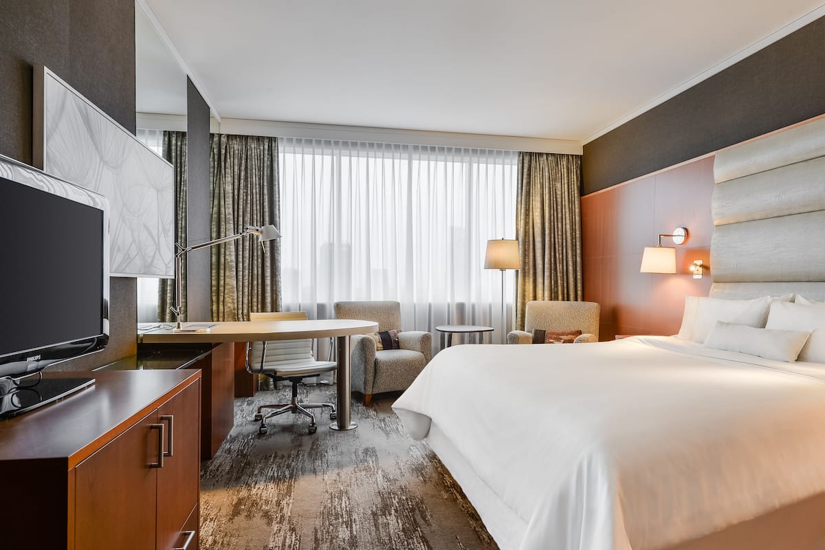 Ein Hotelzimmer mit Bett, Schreibtisch, Stuhl, Fernseher und Blick auf die Stadt durch große Fenster.
