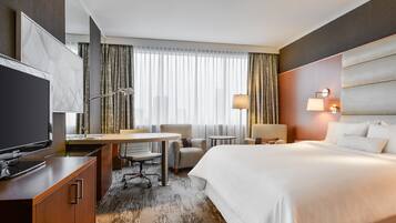 A hotel room with a bed, desk, chair, TV, and a view of the city through large windows.