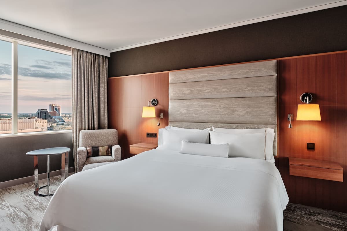 Ein Hotelzimmer mit einem großen Bett, zwei Nachttischen mit Lampen, einem Sessel und Blick auf die Stadt durch große Fenster.