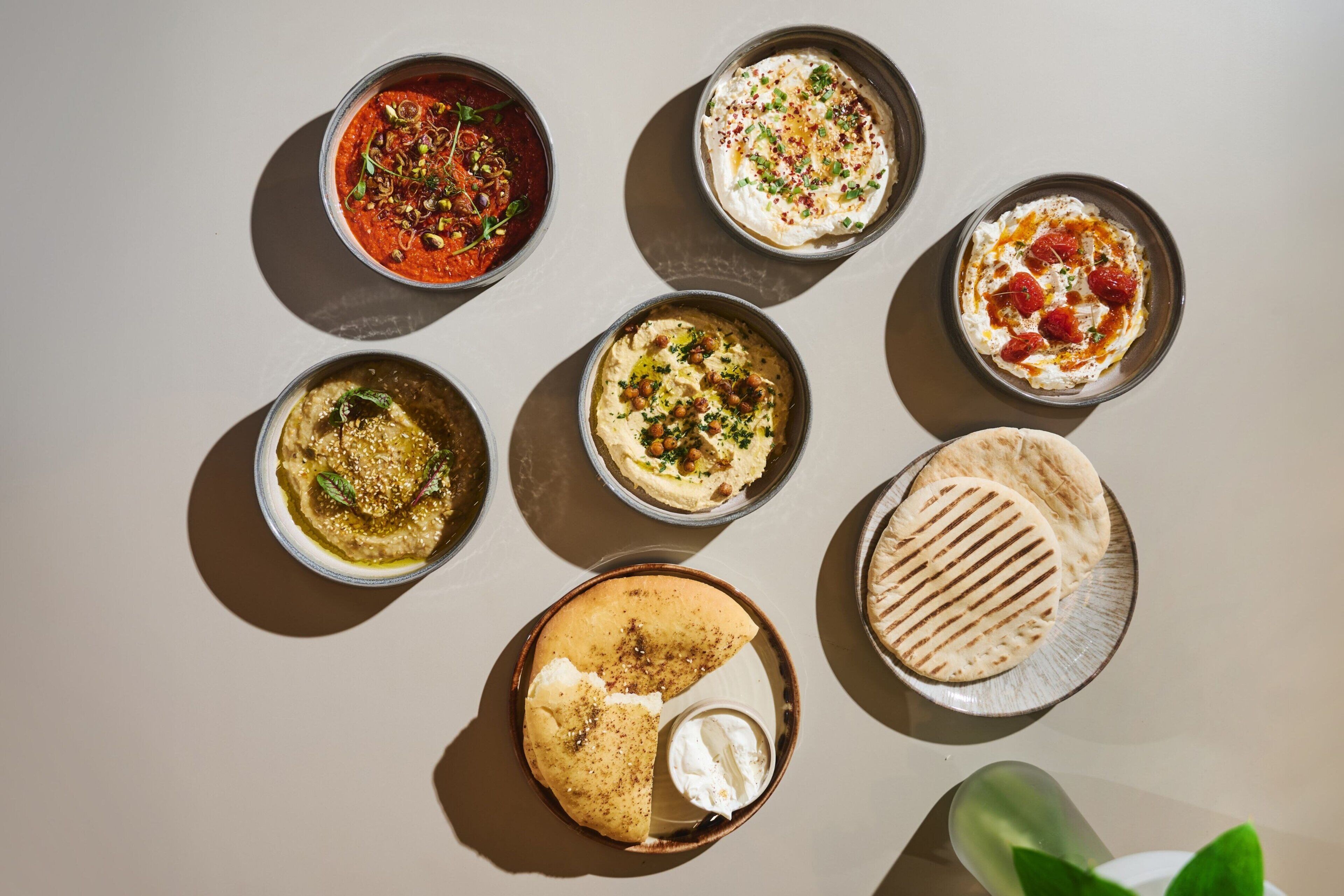 A top-down view of various food items, including bowls of hummus and a grilled flatbread, arranged on a table.