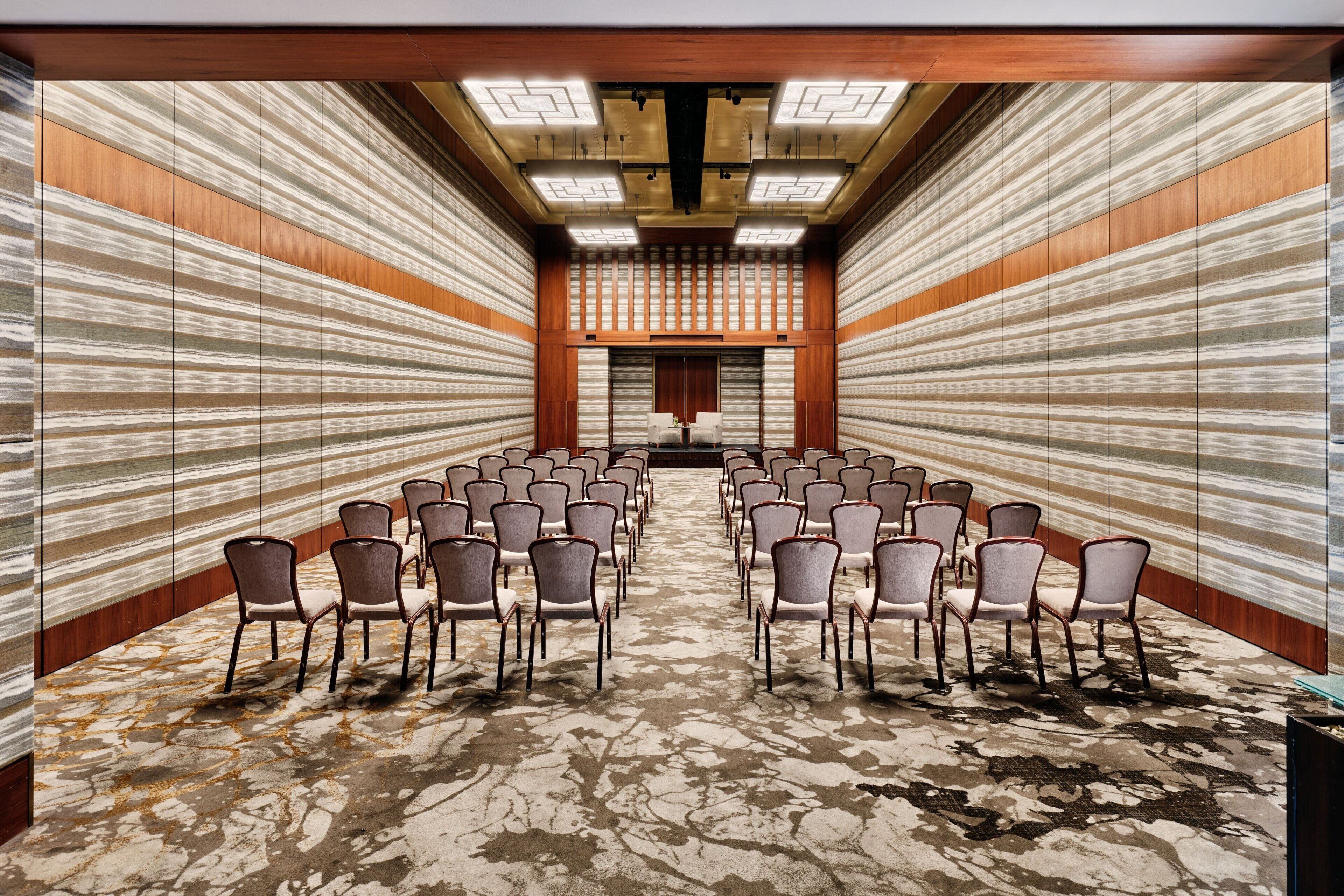 A conference room with rows of chairs facing a podium, wooden panel walls, and a patterned carpet.
