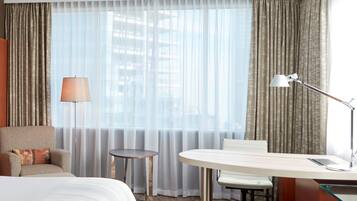 A hotel room with a bed, a desk, a chair, a lamp, and a view of the city through sheer curtains.