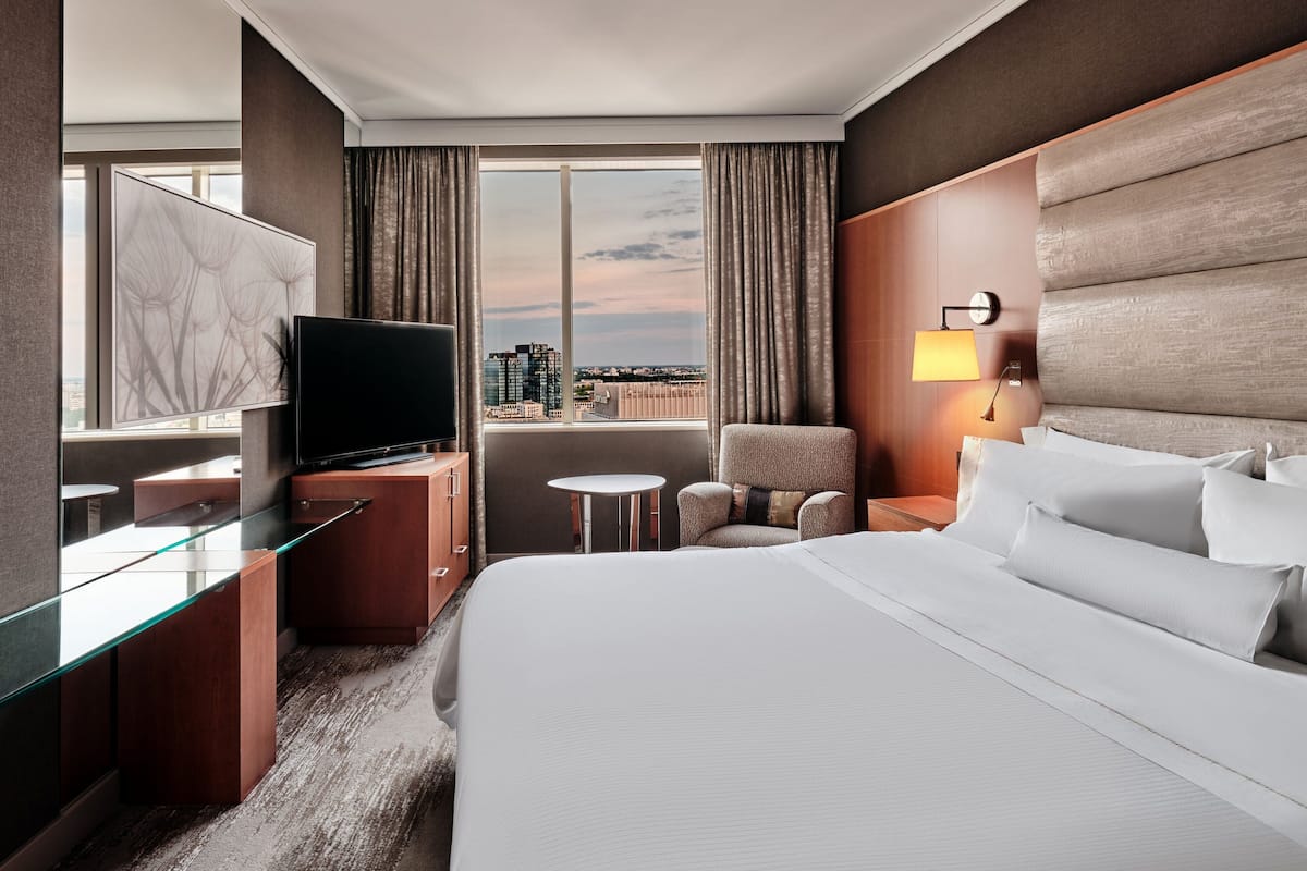 Ein Hotelzimmer mit einem großen Bett, einem Fernseher auf einer Holzkommode, einem kleinen Tisch, einem Sessel und Blick auf die Stadt durch große Fenster.