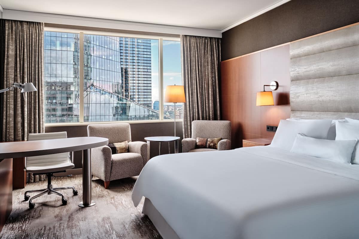 Ein Hotelzimmer mit einem großen Bett, zwei Sesseln, einem kleinen runden Tisch und einem Schreibtisch. Ein Fenster bietet Blick auf die Stadt, und eine Treppe ist ebenfalls zu sehen.