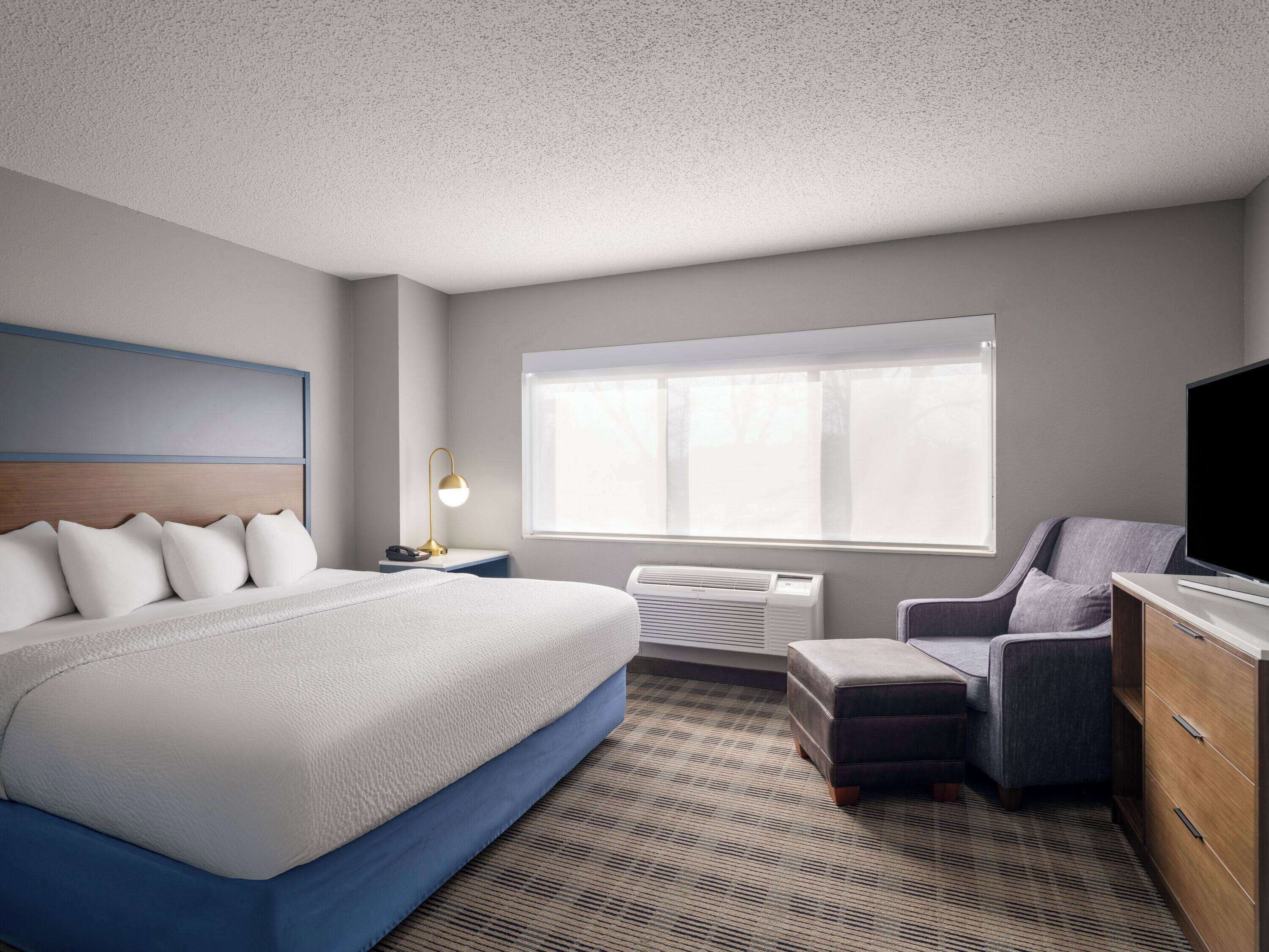 A hotel room with a large bed, a desk with a lamp, a chair, a TV, and a window with blinds.