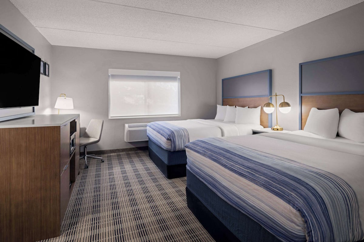 A hotel room with a large bed, a smaller bed, a desk with a TV, a chair, and a window with blinds.