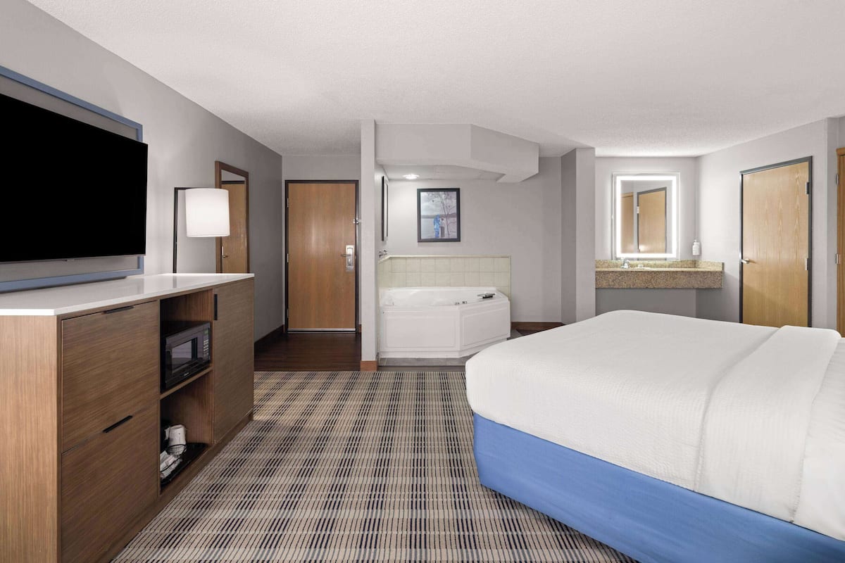 A hotel room with a large bed, a flat-screen TV, a bathroom with a bathtub, and a minibar.