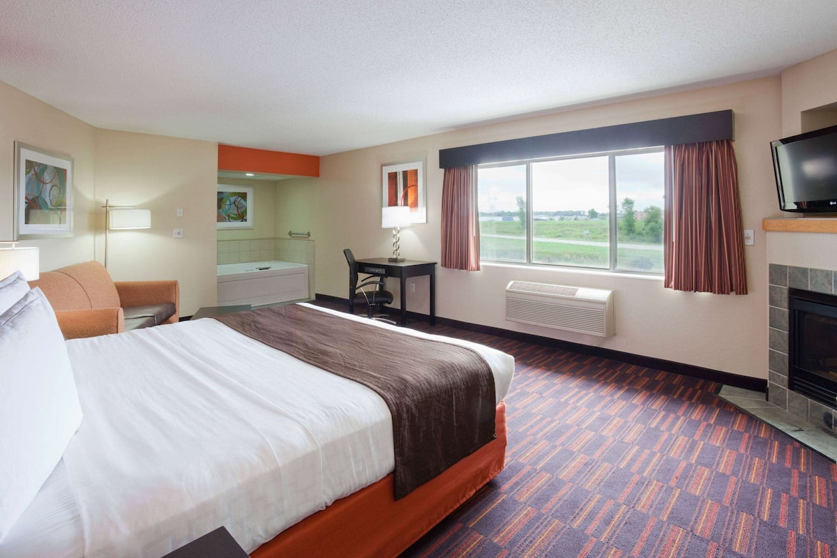 A hotel room with a large bed, a desk, a chair, a television, a fireplace, and a window with curtains.