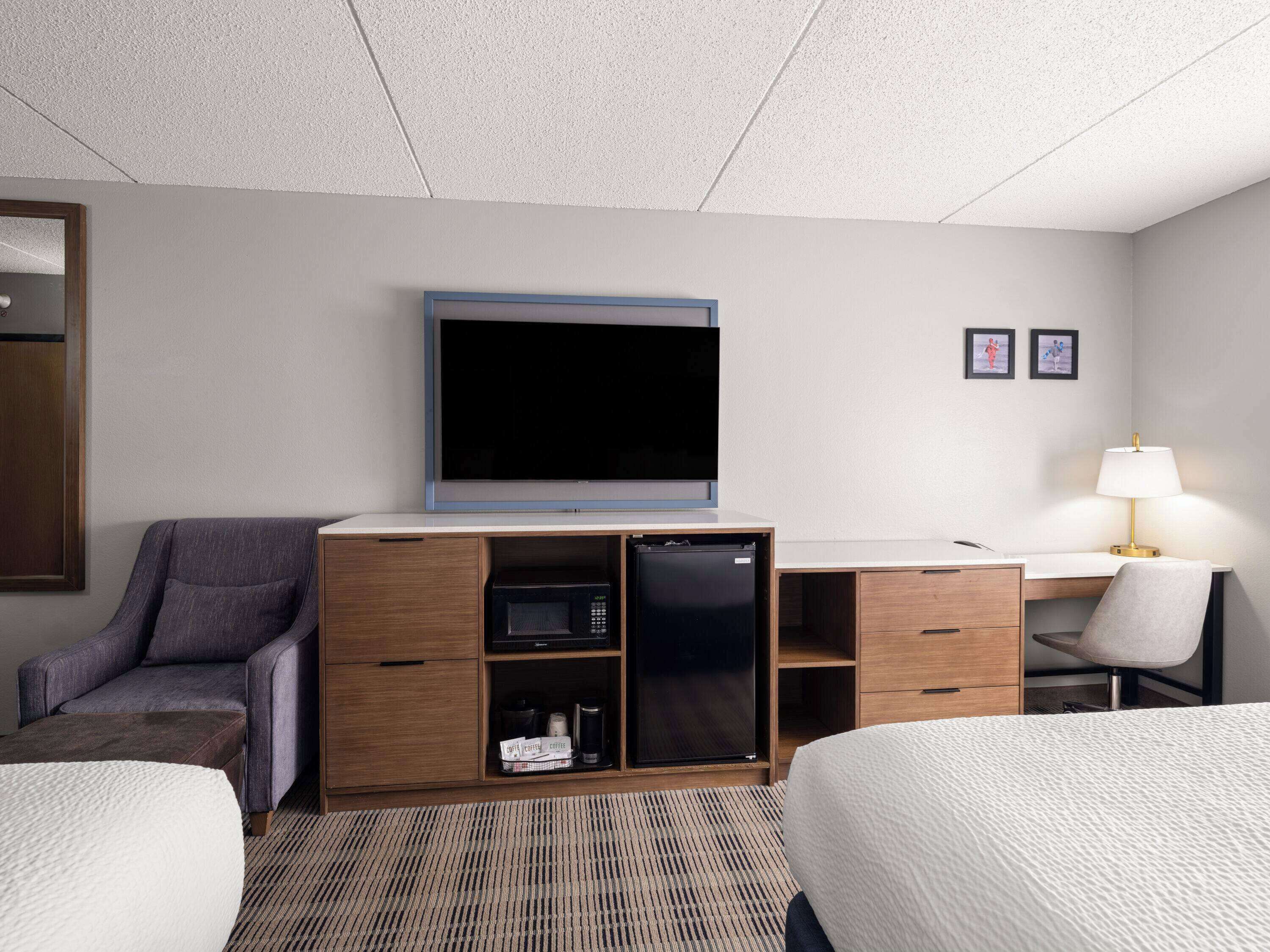 Ein Hotelzimmer mit Bett, Schreibtisch mit Lampe, Fernseher, Minibar und einer Sitzecke mit einem Sessel.