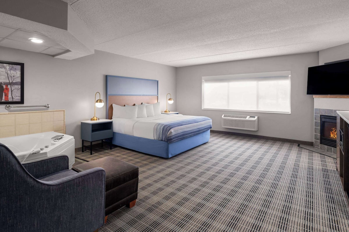 A modern hotel room with a large bed, bedside tables, a flat-screen TV, a fireplace, and a large window.