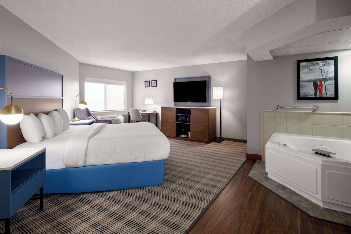 A modern hotel room with a large bed, a desk, a flat-screen TV, a desk lamp, a microwave, a small cabinet, a bathroom with a bathtub, and a large window.