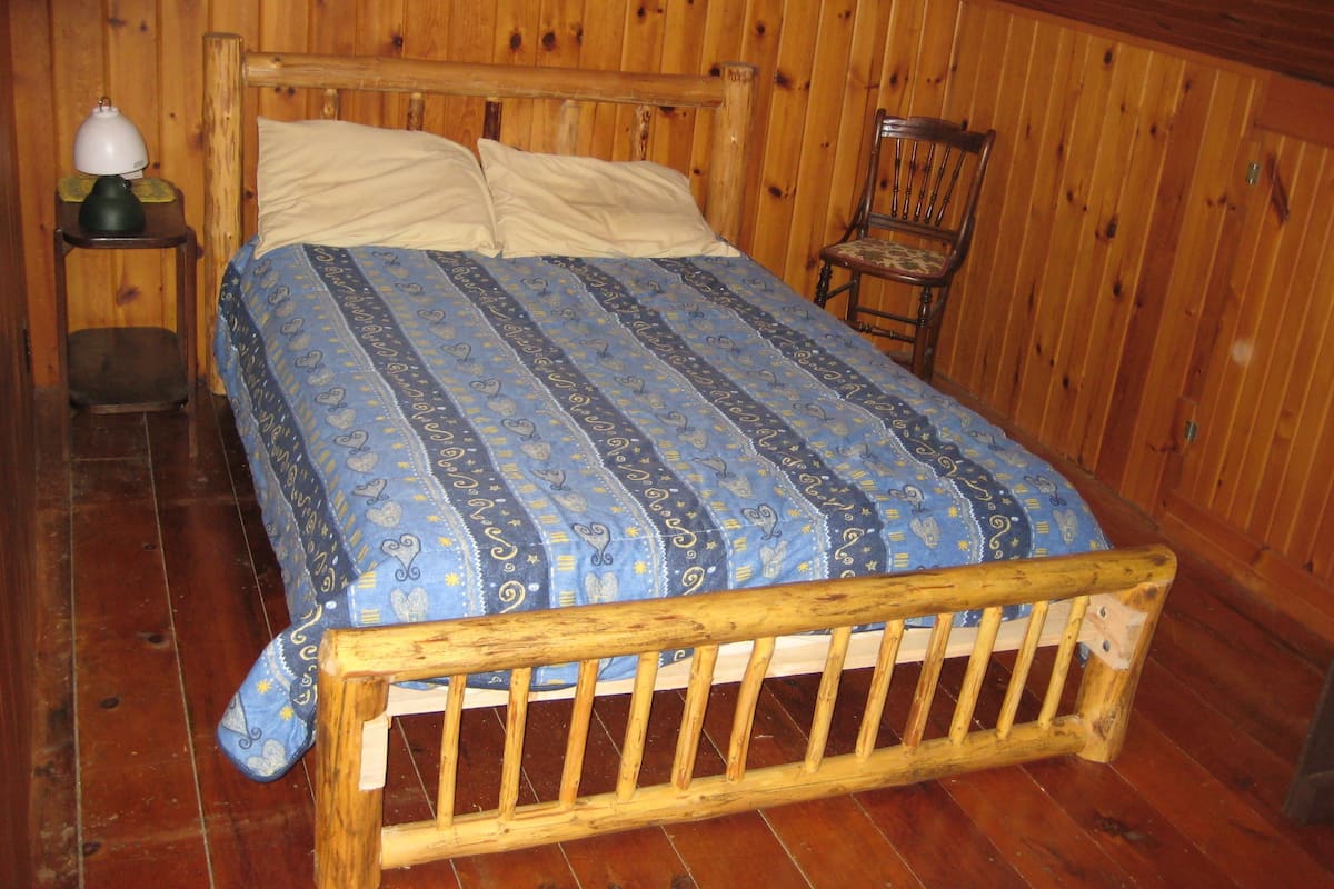 Ein Bett mit Holzrahmen und einem gemusterten Überwurf, ein Nachttisch mit Lampe sowie ein Stuhl in einem Raum mit Holzwänden und Holzboden.