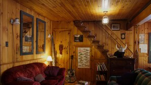 Interior acogedor de una cabaña con paredes de madera, un sofá de terciopelo rojo, un sofá de cuadros verdes, una mesa de centro de madera y una escalera con barandilla de madera.