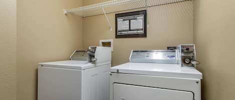 A laundry area with two washers and a dryer, a metal shelf above, and a notice on the wall.