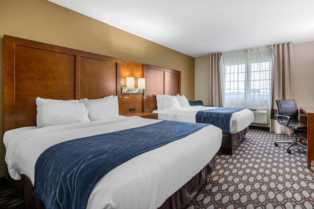 A hotel room with two beds, a desk, a chair, a lamp, and a window with curtains.