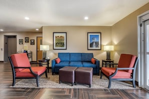 A hotel lobby with a blue sofa, red chairs, a coffee table, two lamps, framed pictures on the wall, and a patterned rug.