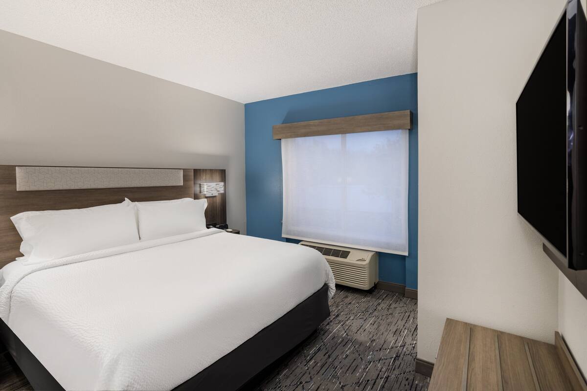 Ein Hotelzimmer mit Bett, Fernseher, einem Fenster mit Jalousien und einer Wand-Klimaanlage.