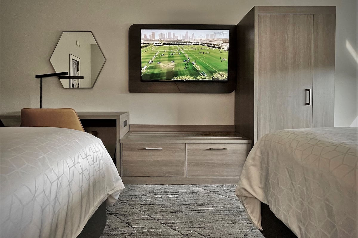 Ein Hotelzimmer mit zwei Betten, einem an der Wand befestigten Fernseher, einem Schreibtisch und einem Spiegel.