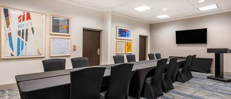 A conference room with a long table, black chairs, a carpeted floor, and a TV on the wall.