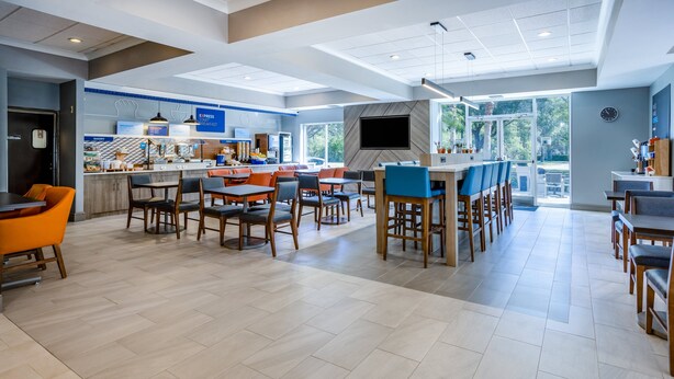 A spacious hotel dining area with multiple seating options, a kitchen area, and a large window providing natural light.