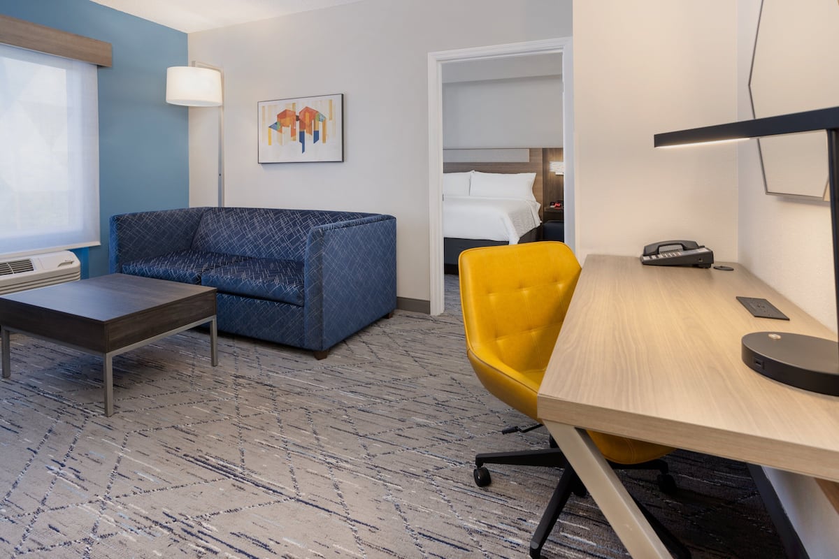 Ein Hotelzimmer mit einem blau gemusterten Sofa, einem gelben Sessel, einem Holztisch und einem Bett im Hintergrund.