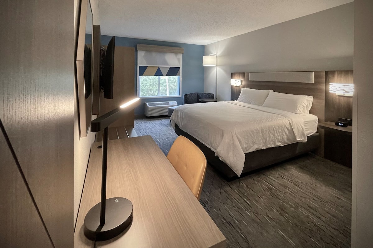 Ein modernes Hotelzimmer mit einem großen Bett, einem Schreibtisch, einem Sessel, einem Flachbildfernseher und einem Fenster mit Blick auf Bäume.