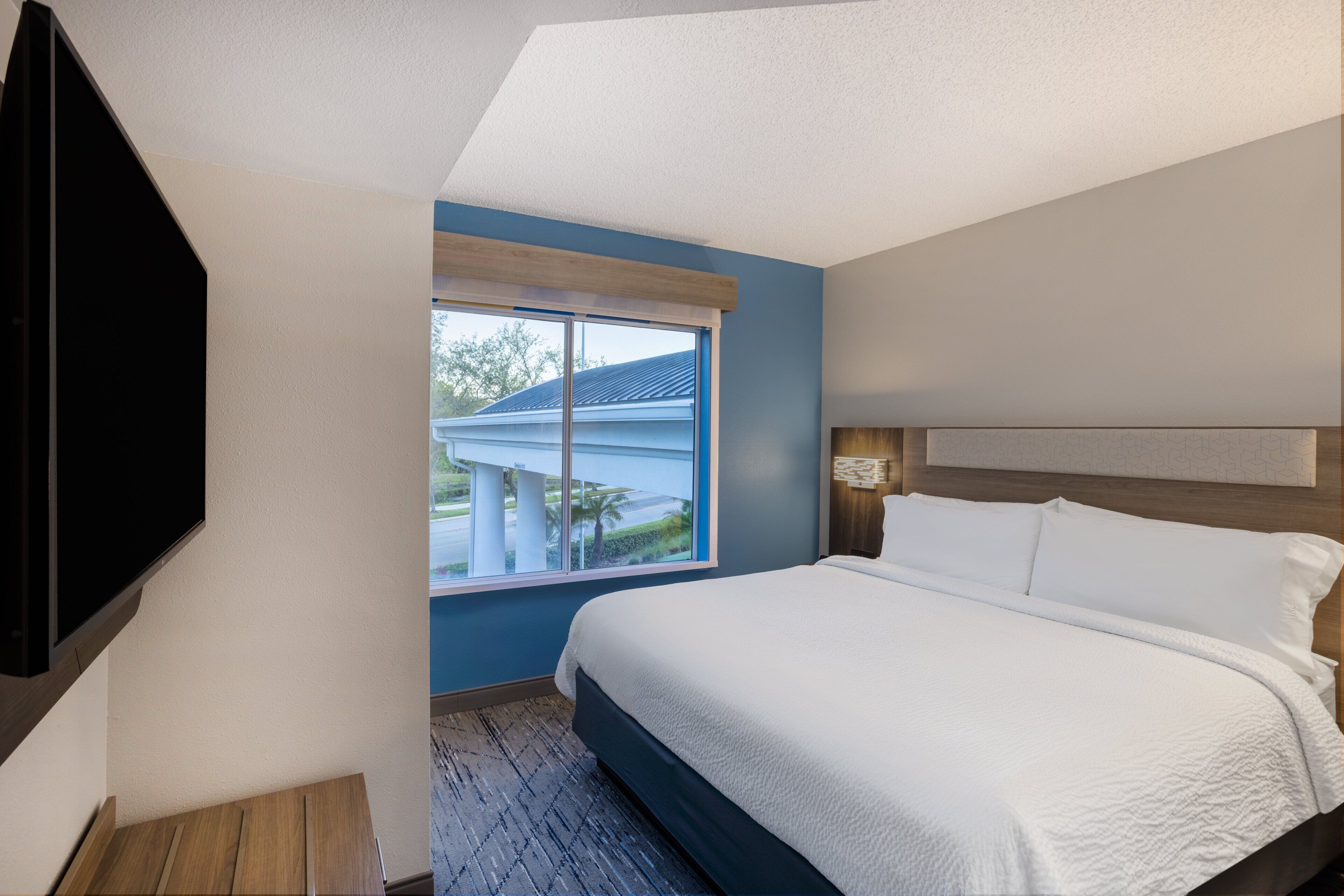 A modern hotel room with a large bed, a flat-screen TV mounted on the wall, and a window offering a view of the outside.