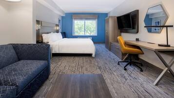 A hotel room with a bed, a sofa, a desk, a chair, a TV, and a window with a view of trees.