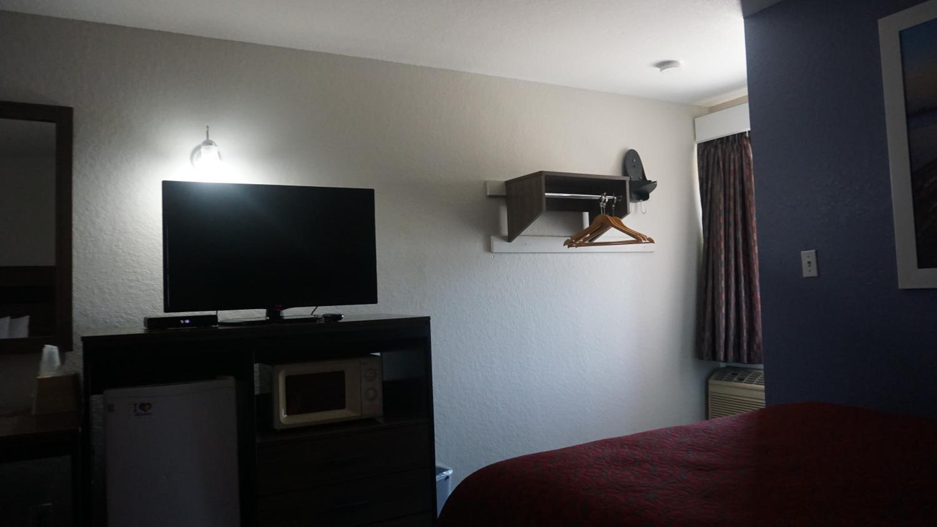 Quarto de hotel com televisão em suporte, micro-ondas, mini-frigorífico, cama com manta vermelha e aquecedor fixado na parede.
