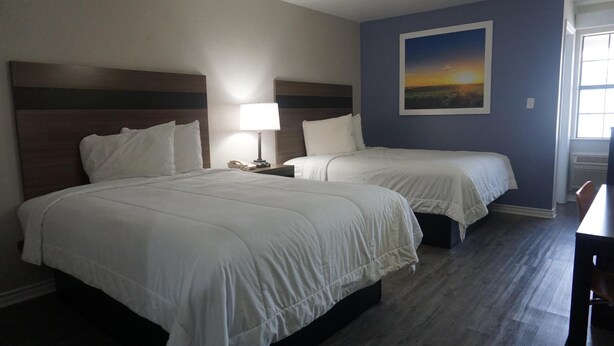 A hotel room with two beds, a nightstand with a lamp, a painting on the wall, and a window. - La Villita Inn (San Antonio)
