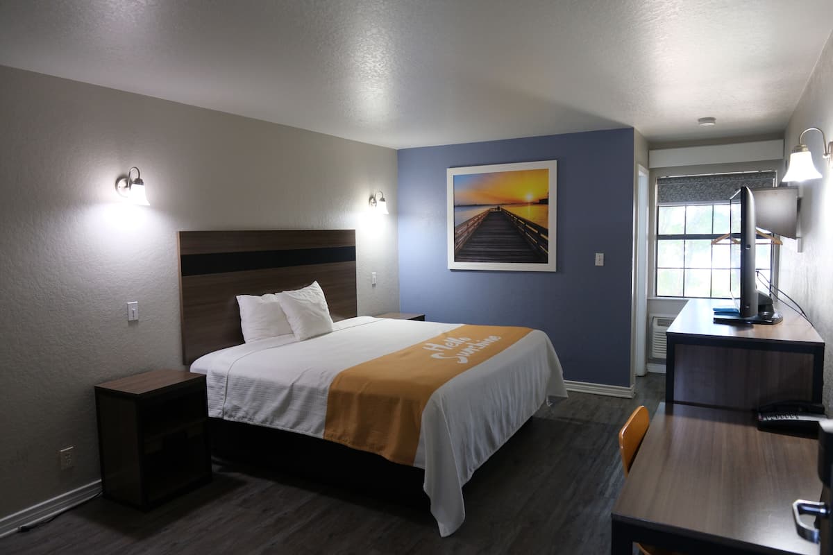 A hotel room with a bed, bedside table, desk, chair, and a painting on the wall.