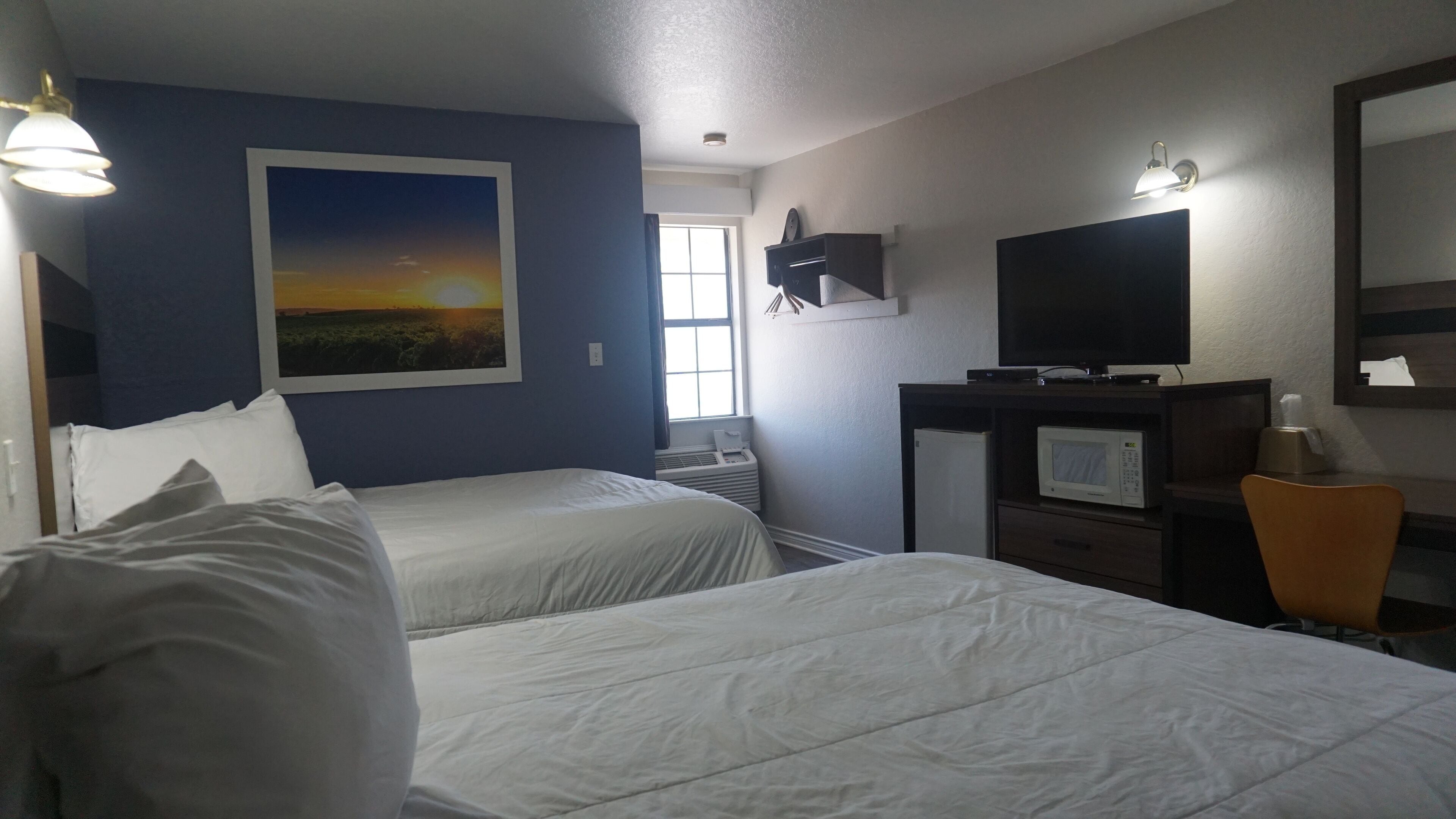 A hotel room with two beds, a TV, a microwave, and a window with a view of the sunset.