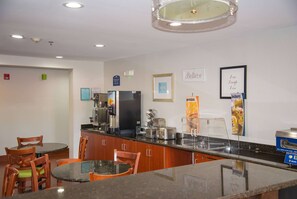 A hotel kitchen with a self-serve breakfast bar, wooden chairs, and a refrigerator.