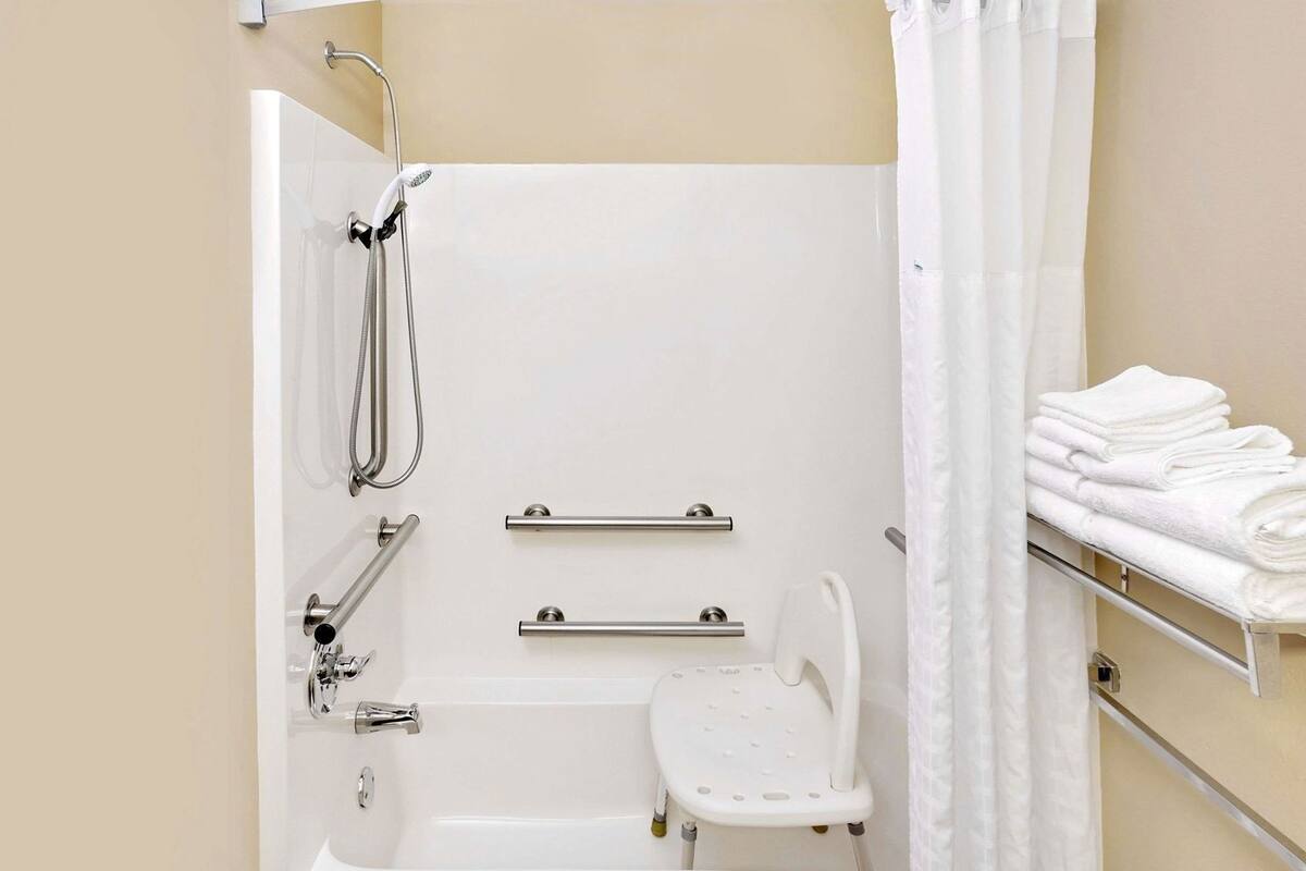 A bathroom with a bathtub, grab bars, a showerhead, a white shower seat, and a towel rack with folded towels.