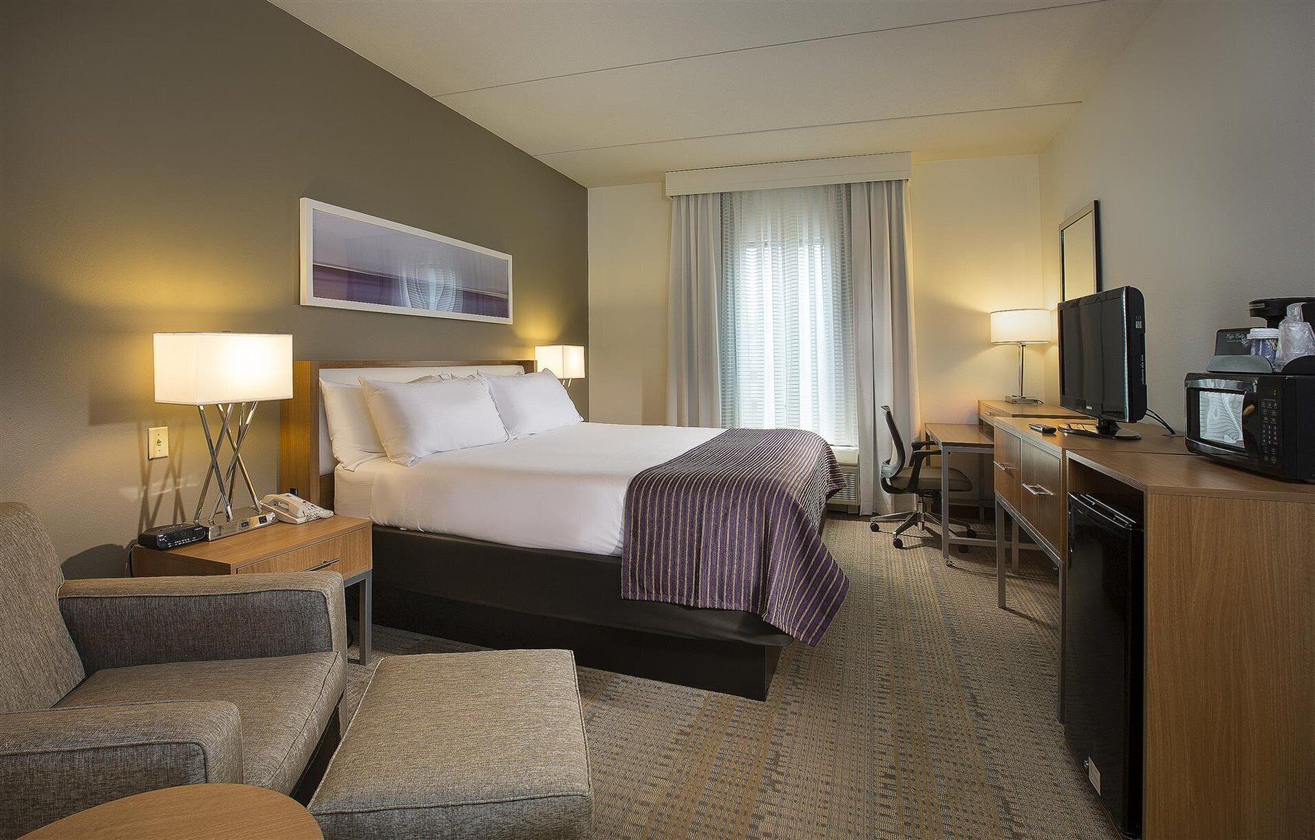Une chambre d’hôtel comprenant un lit, des lampes de chevet, un bureau avec une télévision, un micro-ondes et une fenêtre avec des rideaux.