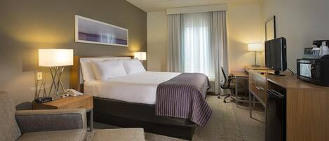 A hotel room with a bed, bedside lamps, a desk with a television, a microwave, and a window with curtains.