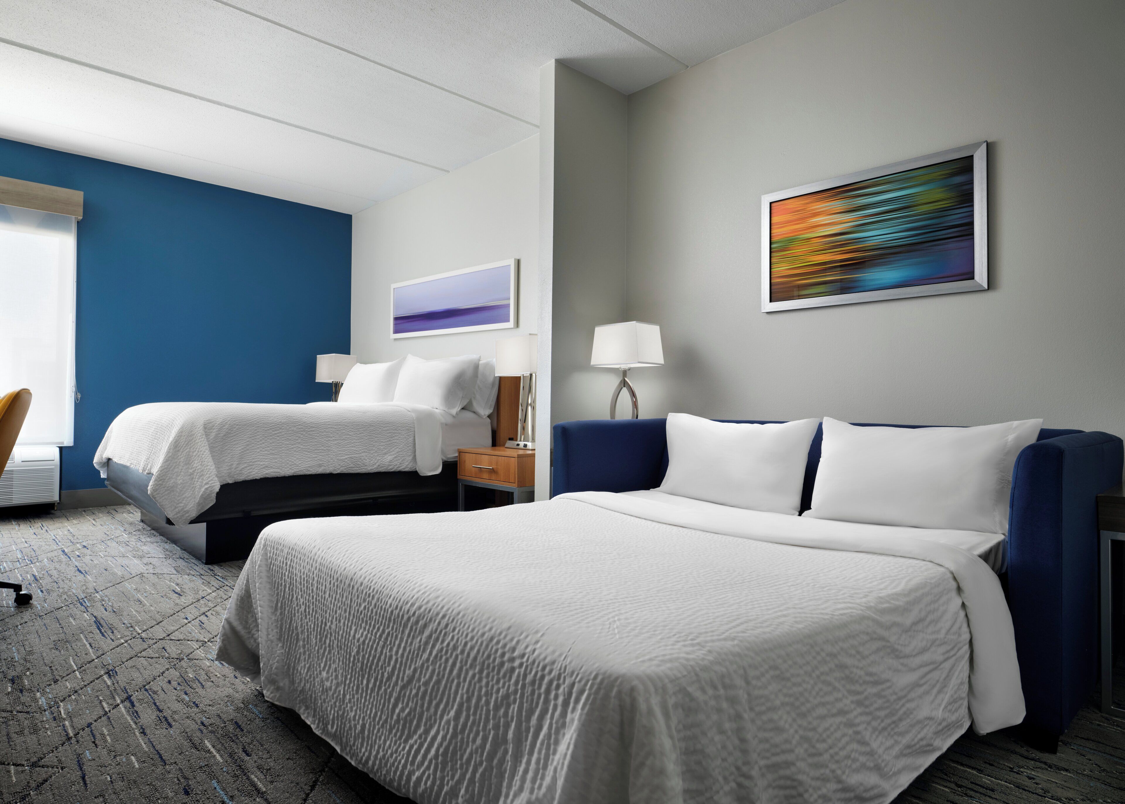 A hotel room with two beds, a desk, a chair, a lamp, and a painting on the wall.