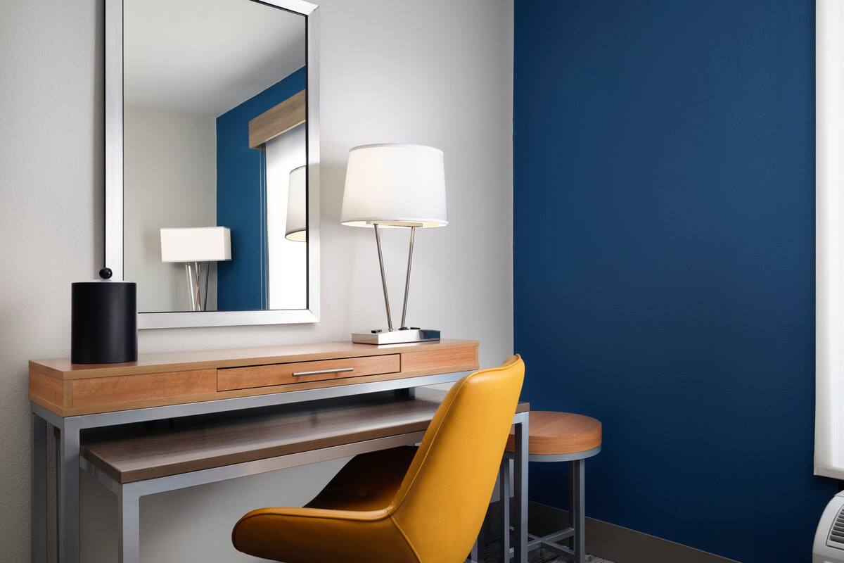 Una habitación de hotel moderna con un escritorio de madera, una silla amarilla, una lámpara blanca y un espejo grande.