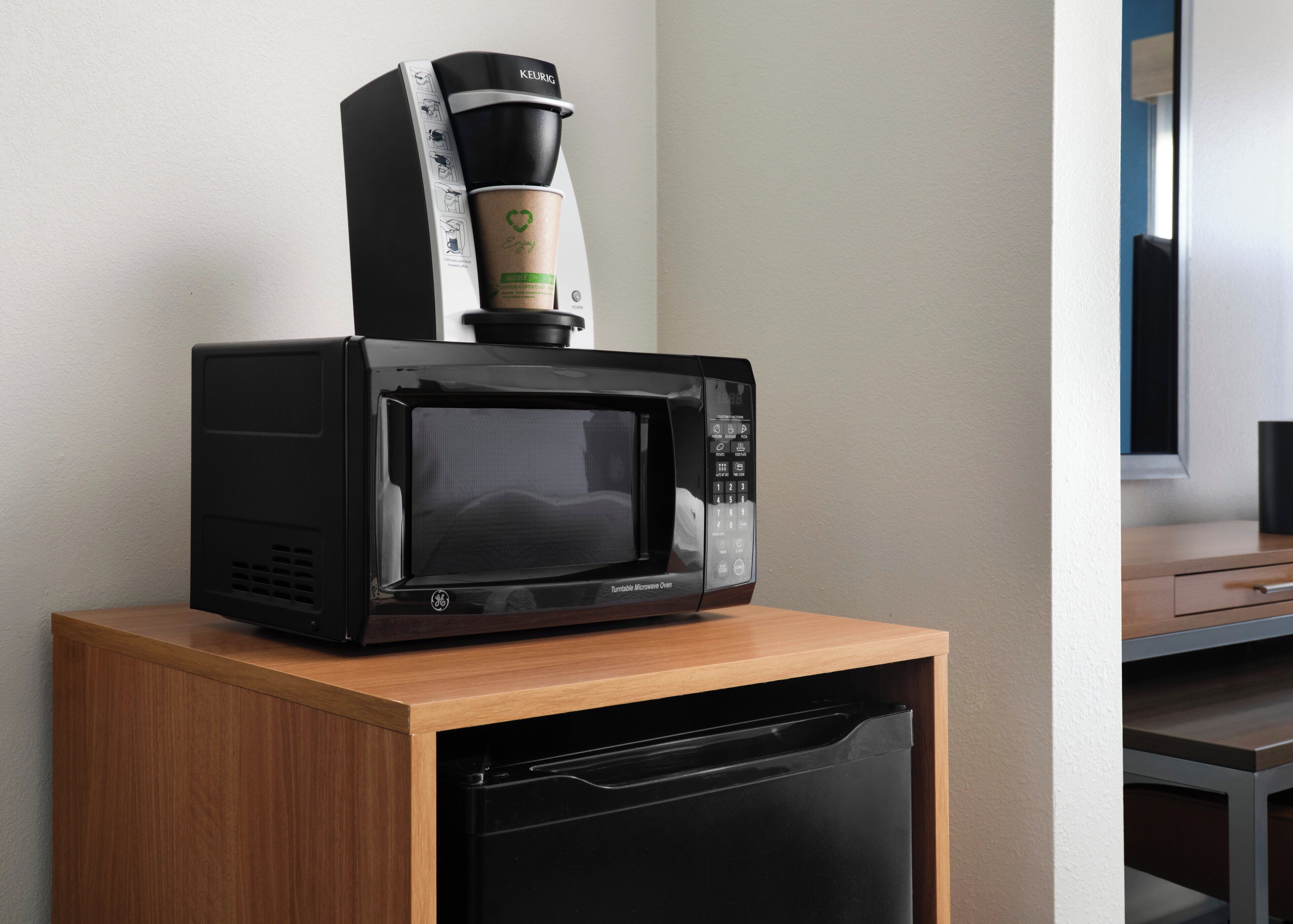 A compact microwave with a coffee maker on top, placed on a wooden cabinet with a dishwasher below.