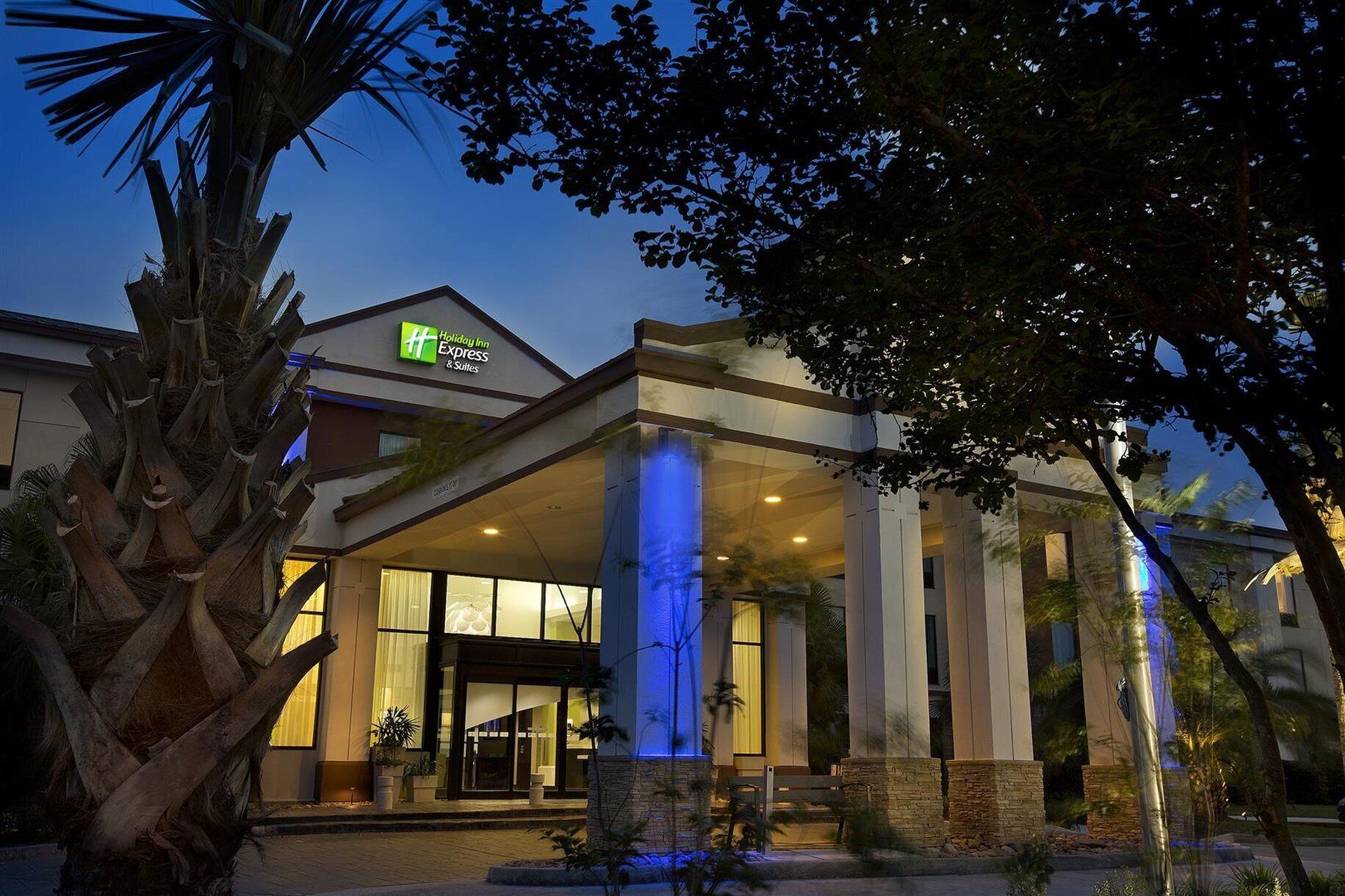 A Holiday Inn Express & Suites hotel with a modern design, featuring a prominent entrance with glass doors and columns, and a well-lit facade.