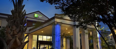 A Holiday Inn Express & Suites hotel with a modern design, featuring a prominent entrance with glass doors and columns, and a well-lit facade.