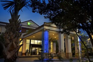 Un hotel Holiday Inn Express & Suites con un diseño moderno, que presenta una entrada destacada con puertas de vidrio y columnas, y una fachada bien iluminada.