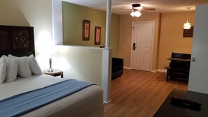 Ein Schlafzimmer mit Bett, Nachttisch, Lampe und einer Tür zu einem Wohnbereich mit Sofa und Klavier.