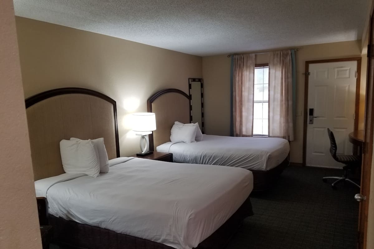 A hotel room with two beds, a nightstand with a lamp, a chair, and a door.