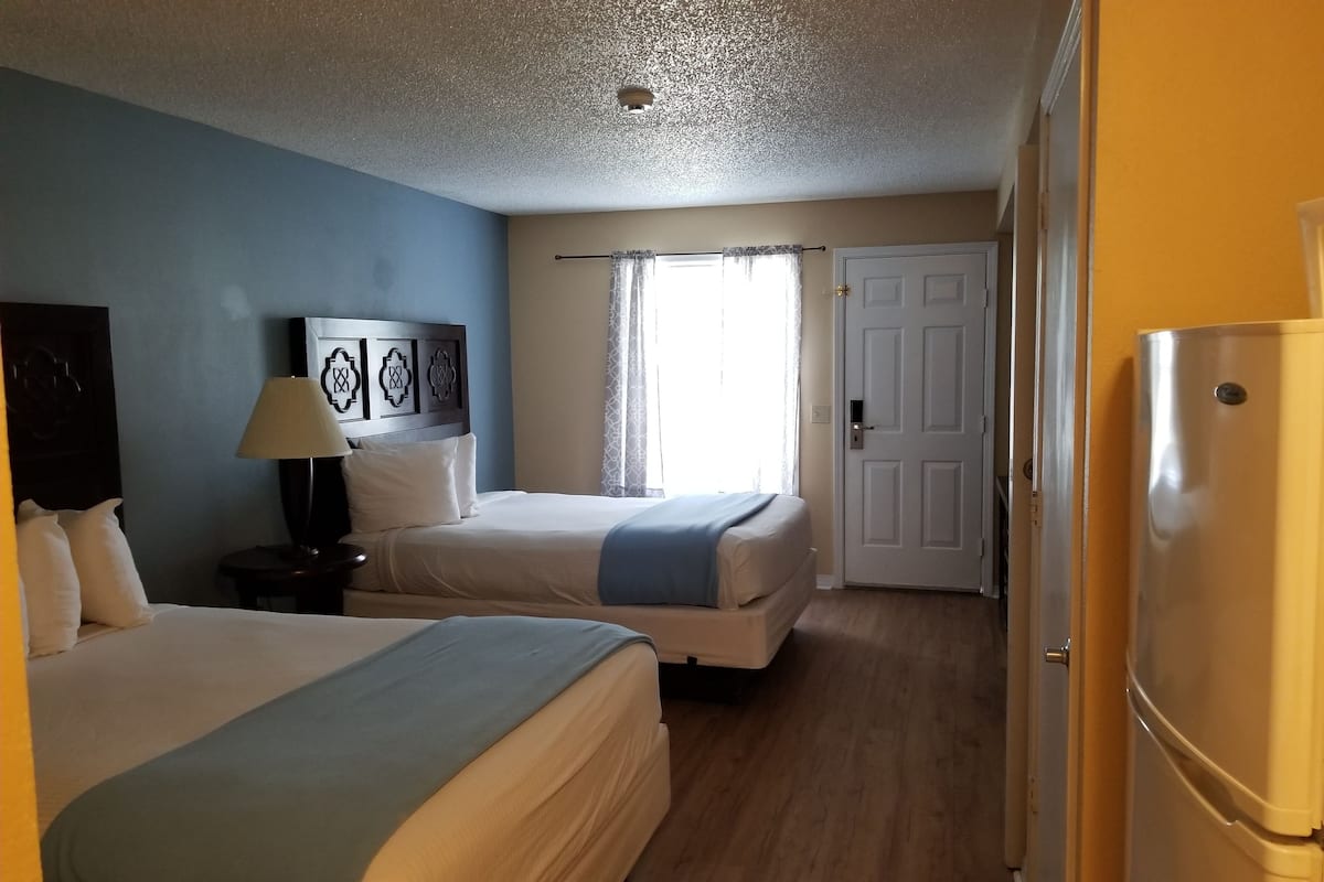A hotel room with two beds, a refrigerator, a nightstand, a lamp, and a door.