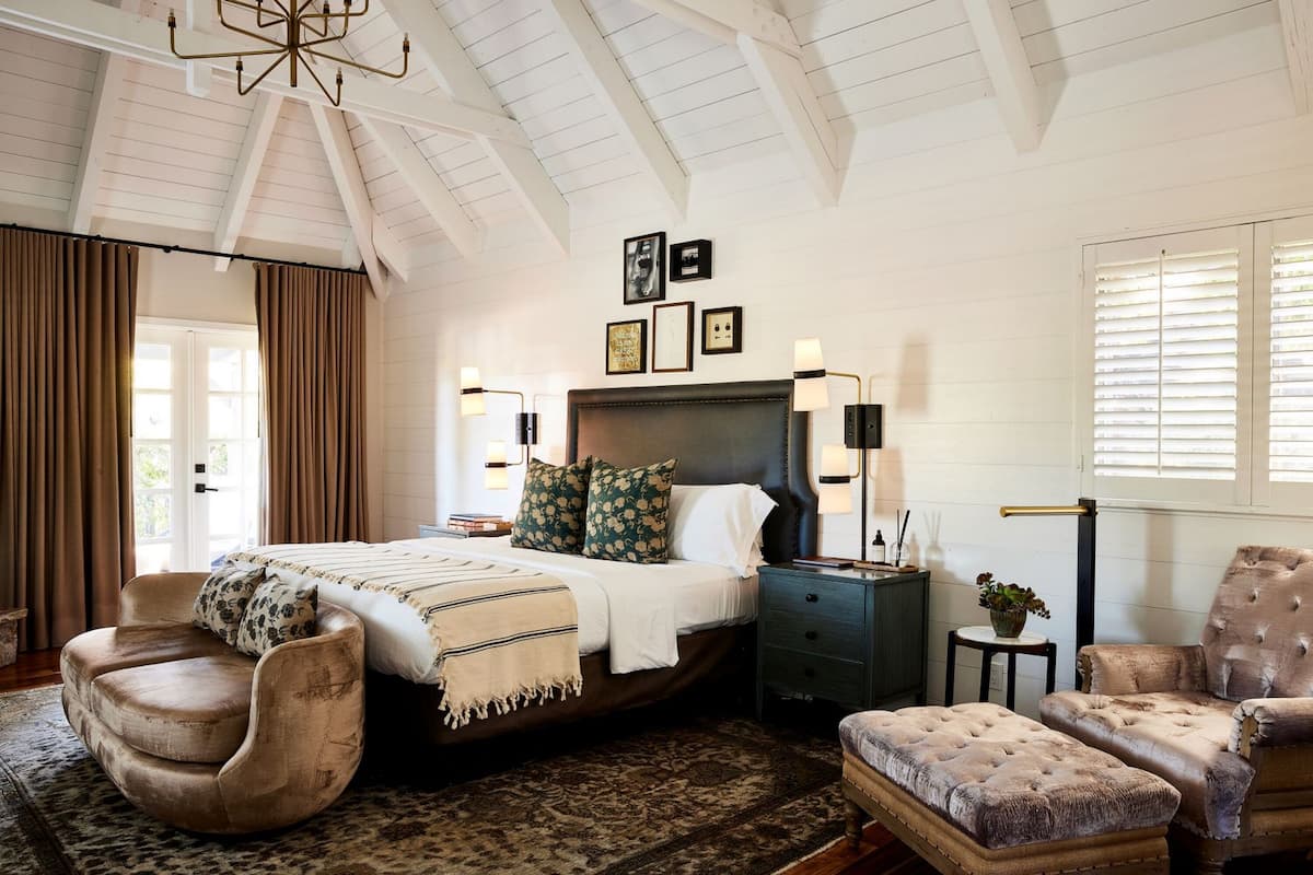 A bedroom with a large bed, a curved armchair, a tufted ottoman, a nightstand with a lamp, and a window with blinds.